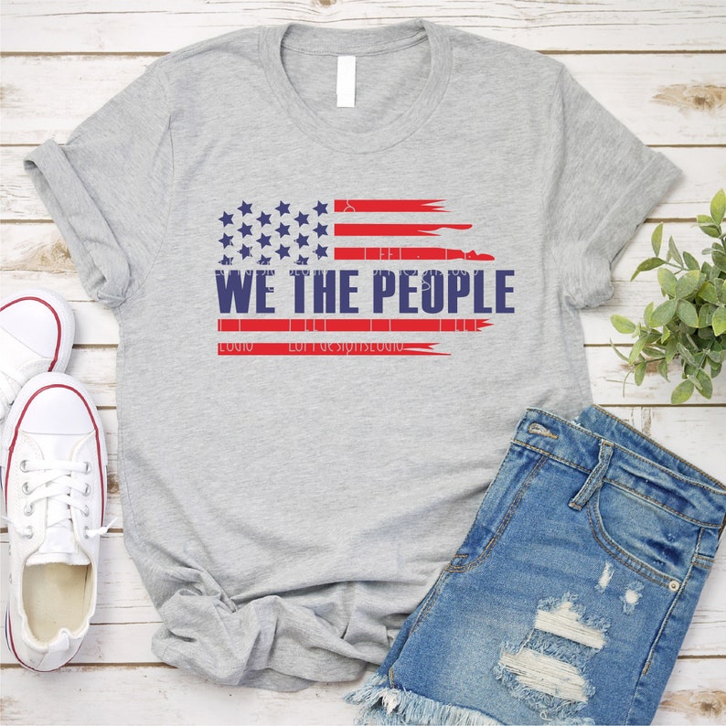 Op de afbeelding: Lichtgrijs t-shirt met een verweerd Amerikaans vlagontwerp met de woorden "WE THE PEOPLE" in marineblauw. Het vlagontwerp bevat rode strepen en blauwe sterren. Het shirt wordt getoond met een spijkerbroekshort en witte sneakers.