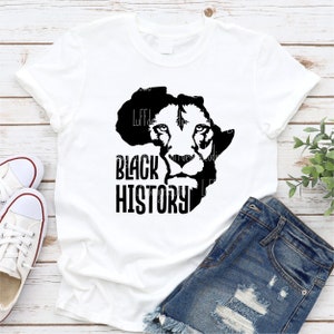 May include: White t-shirt featuring a black graphic of a lion's face within the outline of the African continent, with the words "BLACK HISTORY" in bold, black letters. The shirt is paired with denim shorts and white sneakers.