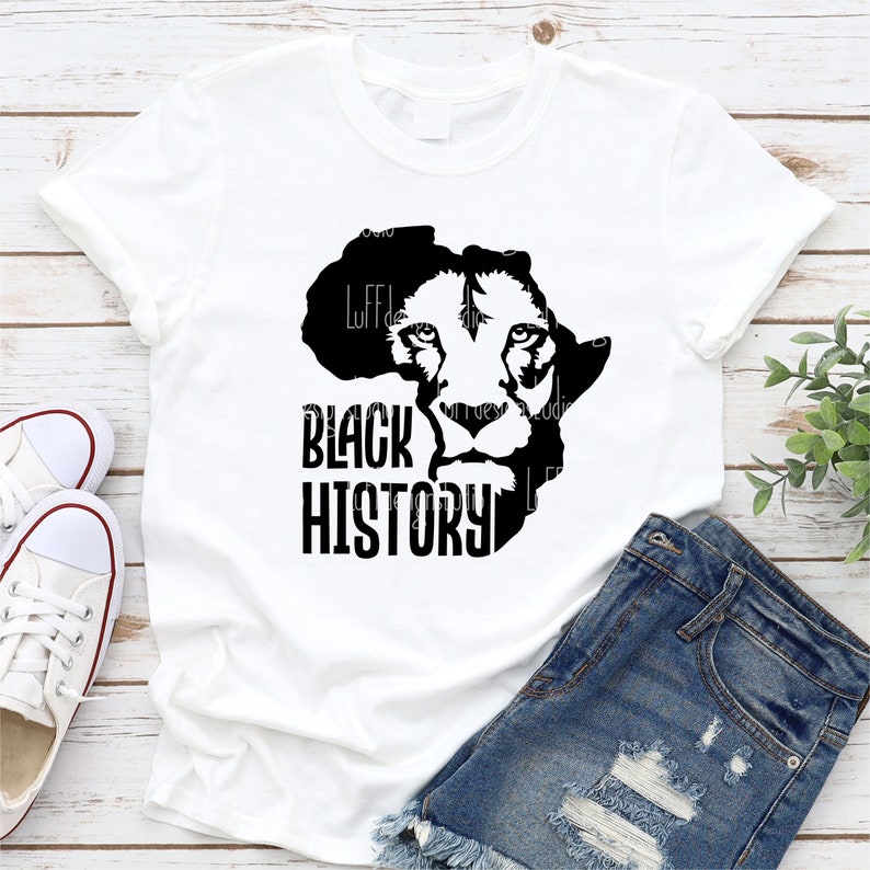 May include: White t-shirt featuring a black graphic of a lion's face within the outline of the African continent, with the words "BLACK HISTORY" in bold, black letters. The shirt is paired with denim shorts and white sneakers.