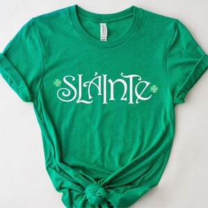 Puede incluir: Camiseta verde con la palabra "Sl&aacute;inte" en letras blancas y dise&ntilde;os de tr&eacute;boles.