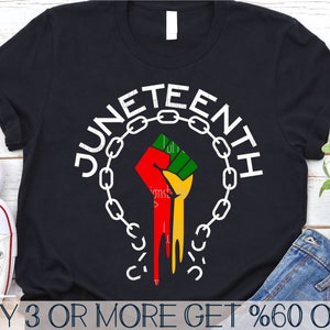Puede incluir: Camiseta negra con un gráfico blanco de un puño levantado liberándose de un círculo de eslabones de cadena. El texto "Juneteenth" está escrito en un arco circular blanco alrededor del puño.