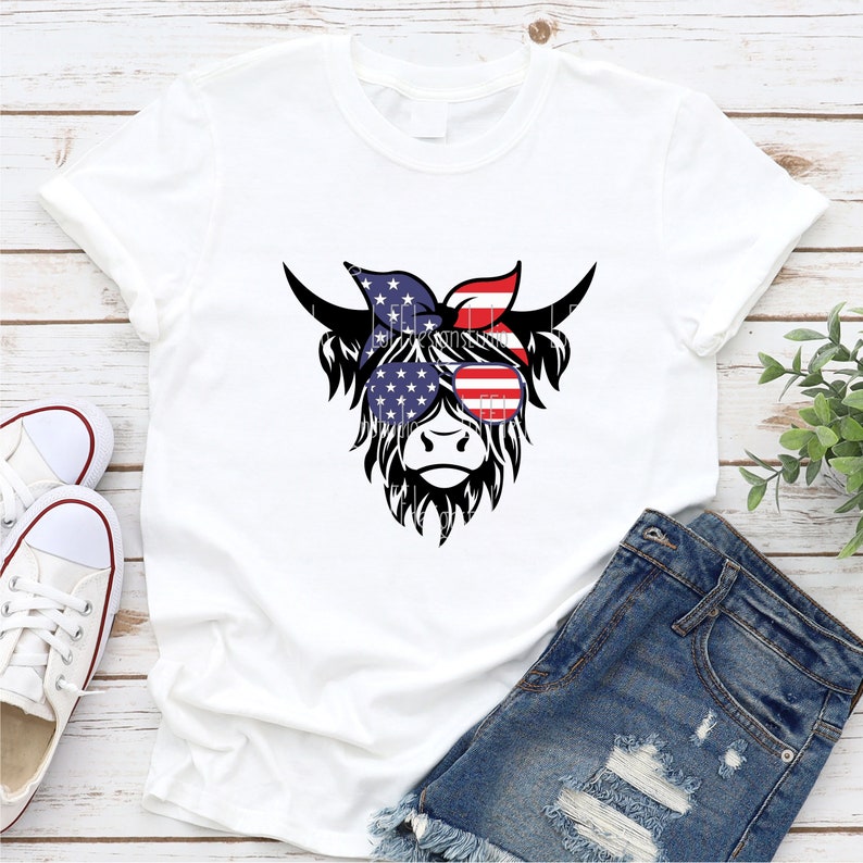 Puede incluir: Una camiseta blanca con una ilustraci&oacute;n en blanco y negro de una vaca de las Tierras Altas con un pa&ntilde;uelo rojo, blanco y azul y gafas de sol. El pa&ntilde;uelo y las gafas de sol est&aacute;n estampados con la bandera estadounidense.