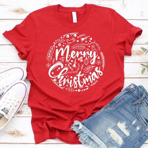 May include: Red t-shirt with a white graphic design of a circle with the words "Merry Christmas" and various Christmas-themed illustrations.