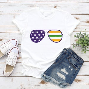 May include: White t-shirt with a graphic of aviator sunglasses. The sunglasses have an American flag design on the left lens and a green, orange, and yellow striped design on the right lens.