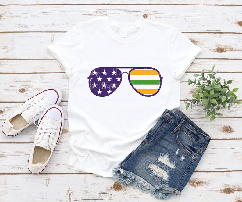 May include: White t-shirt with a graphic of aviator sunglasses. The sunglasses have an American flag design on the left lens and a green, orange, and yellow striped design on the right lens.