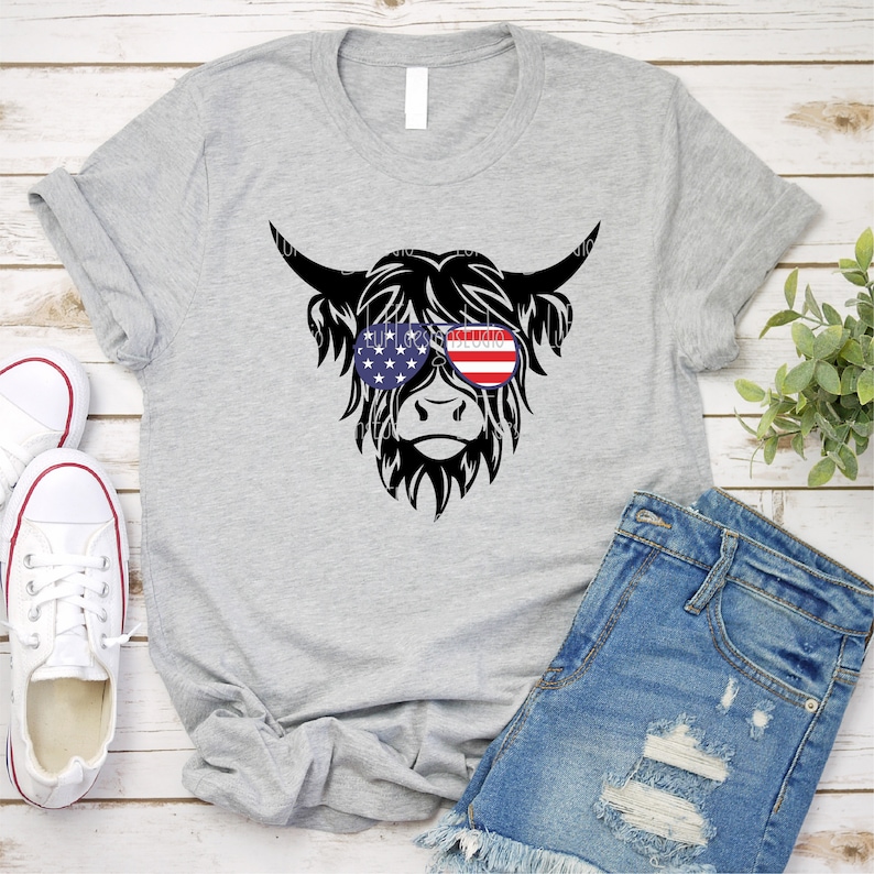 Puede incluir: Una camiseta gris con una vaca de las tierras altas negra que lleva gafas de sol con el dise&ntilde;o de la bandera estadounidense. La vaca mira hacia adelante.