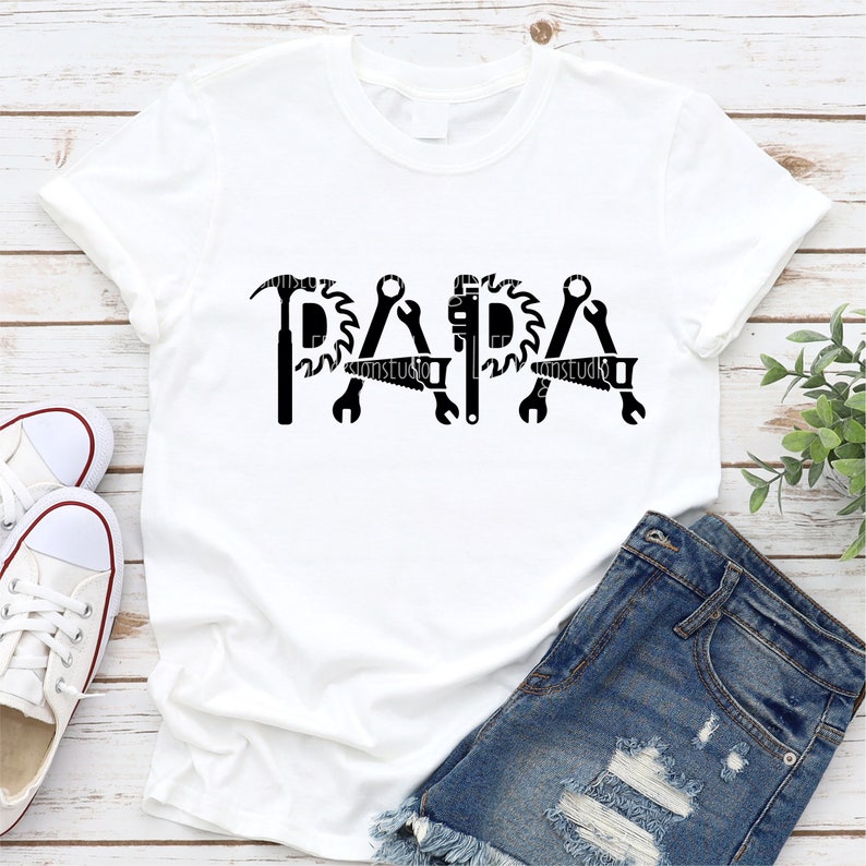 Peut inclure: T-shirt blanc avec un motif noir du mot "PAPA" compos&eacute; d'outils comme un marteau, une cl&eacute;, une scie et des pinces.