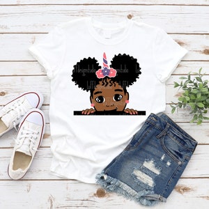 May include: White t-shirt featuring a graphic of a Black girl with pink and purple flowers in her hair and a unicorn horn. The girl is peeking over a surface with her hands on the edge.