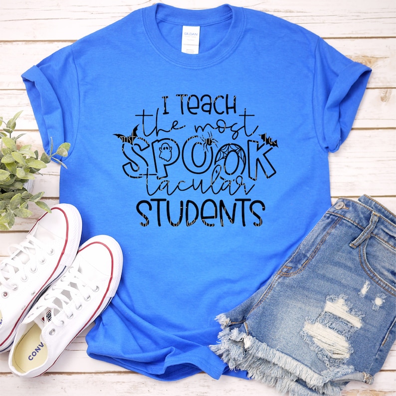 以下が含まれることがあります： ロイヤルブルーのTシャツに、黒い文字で「I teach the most spooktacular students」と書かれています。文字はコウモリ、クモ、クモの巣で飾られています。