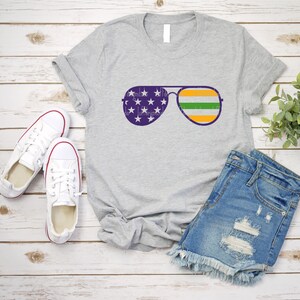 May include: A gray t-shirt with a design of aviator sunglasses with the American flag on the lenses and an Irish flag on the frame. The t-shirt is laid out on a white wooden surface with a pair of white sneakers and a pair of denim shorts.