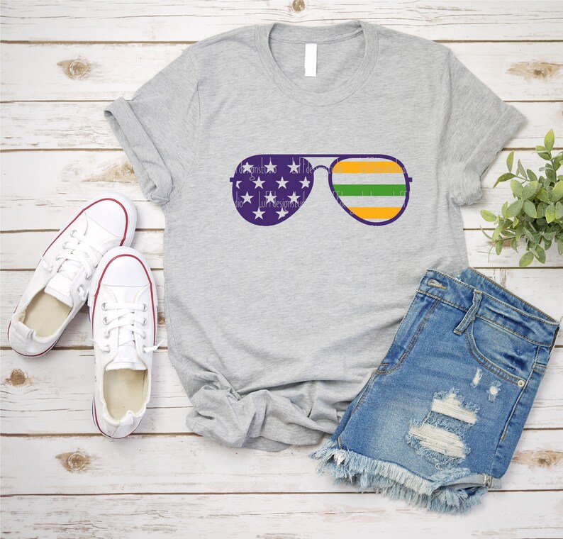 May include: A gray t-shirt with a design of aviator sunglasses with the American flag on the lenses and an Irish flag on the frame. The t-shirt is laid out on a white wooden surface with a pair of white sneakers and a pair of denim shorts.