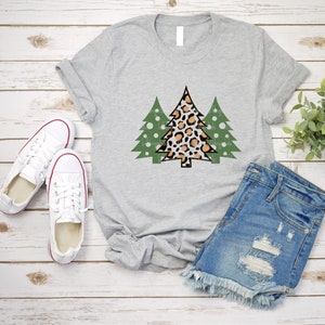 K&ouml;nnte beinhalten: Hellgraues T-Shirt mit einem Weihnachtsbaum-Design. Der zentrale Baum hat ein Leopardenmuster in Braun und Beige, flankiert von zwei gr&uuml;nen B&auml;umen mit wei&szlig;en Punkten. Das Shirt wird mit Jeansshorts und wei&szlig;en Turnschuhen kombiniert.