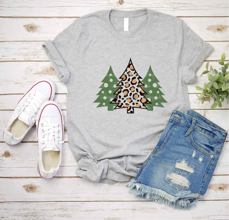 K&ouml;nnte beinhalten: Hellgraues T-Shirt mit einem Weihnachtsbaum-Design. Der zentrale Baum hat ein Leopardenmuster in Braun und Beige, flankiert von zwei gr&uuml;nen B&auml;umen mit wei&szlig;en Punkten. Das Shirt wird mit Jeansshorts und wei&szlig;en Turnschuhen kombiniert.