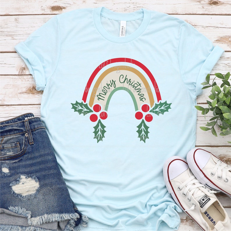 May include: A light blue t-shirt with a rainbow design featuring the text "Merry Christmas" in a vintage style. The rainbow is decorated with holly and red berries.
