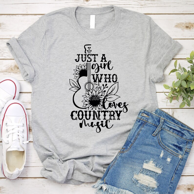 Puede incluir: Camiseta gris claro con el texto negro "JUST A girl WHO loves COUNTRY MUSIC". El dise&ntilde;o presenta una forma de guitarra con girasoles y hojas. La camiseta se muestra con pantalones cortos vaqueros y zapatillas blancas.