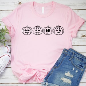 May include: A pink t-shirt with a row of four black and white pumpkin faces with different expressions. The pumpkins are all the same size and shape.