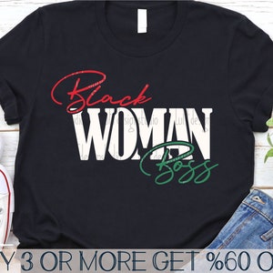 May include: Black t-shirt with white text that reads "Black Woman Boss" in a red, white, and green color scheme. The text is in a stylized font. The t-shirt is on a wooden surface with a pair of jeans and a pair of sneakers.