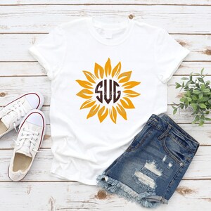 Puede incluir: Camiseta blanca con un monograma marr&oacute;n "SVG" dentro de un gr&aacute;fico de girasol amarillo.