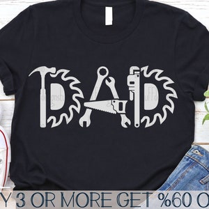 May include: A black t-shirt featuring a white graphic design of the word "DAD" formed from tools such as a hammer, saw, and wrench. The tools are encircled by saw blades.