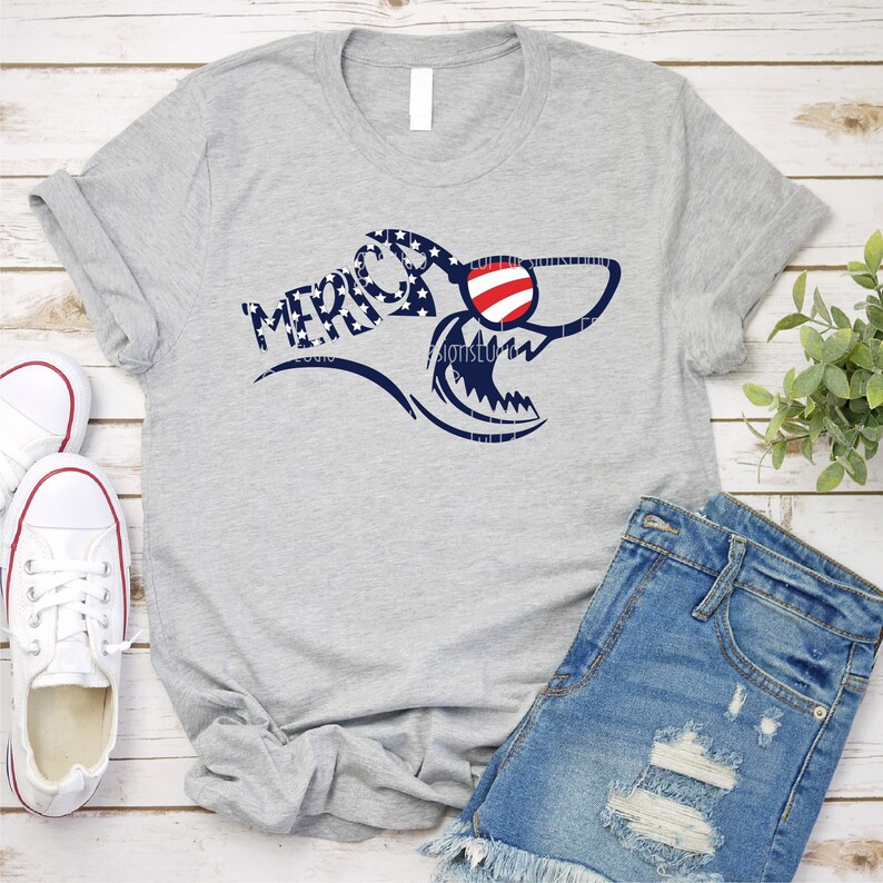 May include: A gray t-shirt with a shark graphic wearing sunglasses. The shark is patterned with the American flag and the word "MERICA" is written on the shark's body.