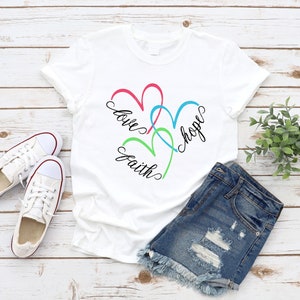 May include: White t-shirt with a design of three intertwined hearts in pink, green, and blue. The words "love", "faith", and "hope" are written in black cursive around the hearts.