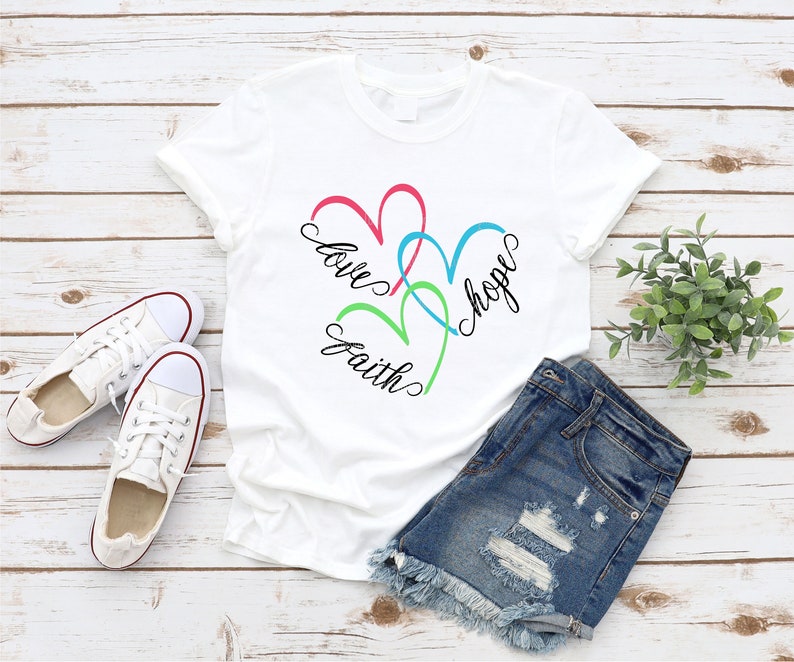 May include: White t-shirt with a design of three intertwined hearts in pink, green, and blue. The words "love", "faith", and "hope" are written in black cursive around the hearts.