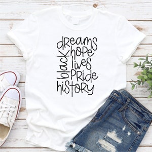May include: White t-shirt with black text that reads "dreams hope black lives pride history".