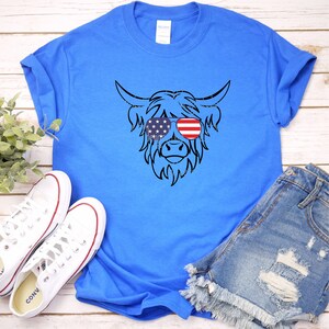 Puede incluir: Una camiseta azul real con una silueta negra de una vaca de las Tierras Altas con gafas de sol con un dise&ntilde;o de bandera estadounidense.