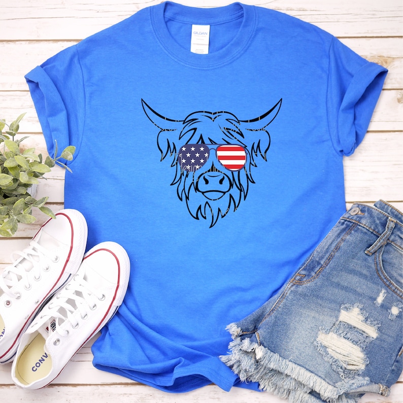 Puede incluir: Una camiseta azul real con una silueta negra de una vaca de las Tierras Altas con gafas de sol con un dise&ntilde;o de bandera estadounidense.