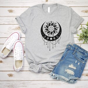 May include: A gray t-shirt with a black and white sun and moon design. The design features a sun with a face and a crescent moon with stars and beads hanging from it.