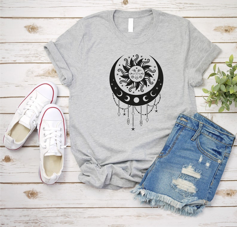 May include: A gray t-shirt with a black and white sun and moon design. The design features a sun with a face and a crescent moon with stars and beads hanging from it.