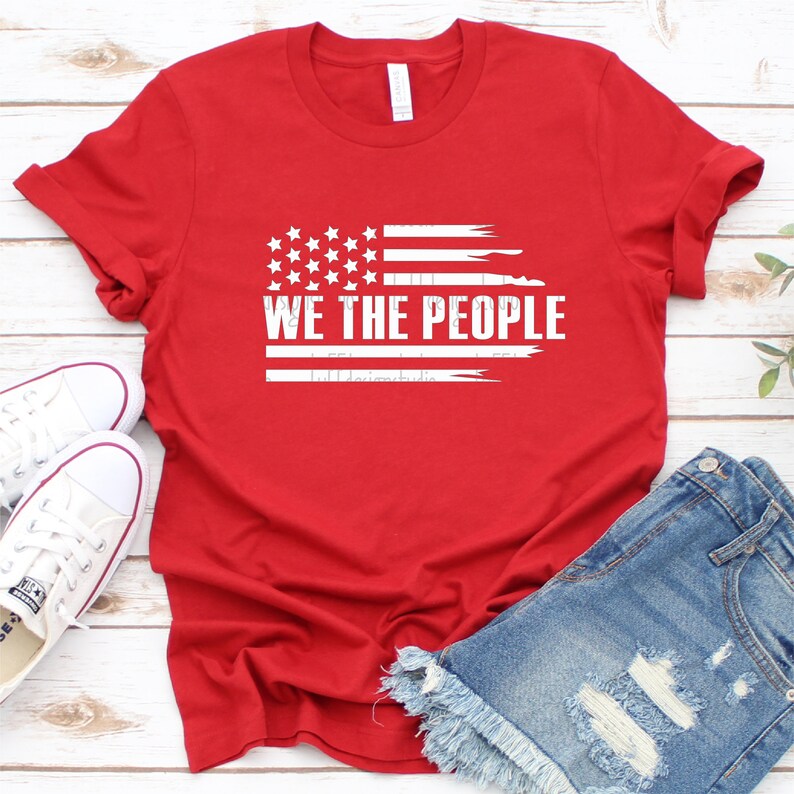 Op de afbeelding: Een rood t-shirt met een versleten Amerikaans vlagontwerp en de witte tekst "WE THE PEOPLE". Het shirt wordt getoond met een spijkerbroek en witte canvas sneakers.