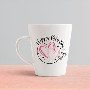 Puede incluir: Taza de cer&aacute;mica blanca con un dise&ntilde;o circular negro con el texto "Happy Valentine's Day" y dos corazones rosas.
