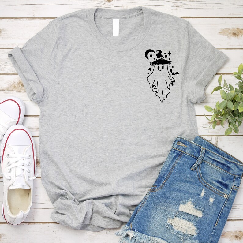 May include: A gray t-shirt with a black graphic of a ghost wearing a witch hat and surrounded by stars. The ghost has a cute, cartoon-like expression.