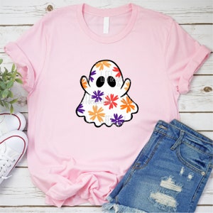 May include: A pink t-shirt with a white ghost design. The ghost is covered in a pattern of orange, purple, and yellow flowers.