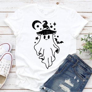 May include: A white t-shirt with a black and white graphic of a ghost wearing a witch hat. The ghost has a smiling face and is surrounded by stars and a crescent moon.