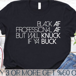 May include: Black t-shirt with white text that reads "Black AF Professional AF But Will Knuck If Ya Buck". The shirt is laying on a wooden surface with a pair of blue jeans and a pair of white sneakers.