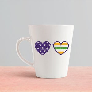 May include: White ceramic mug with a purple and white heart-shaped sunglasses design on one side. The sunglasses have a green, yellow, and purple striped heart on the right lens.
