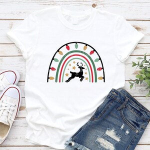 May include: White t-shirt with a rainbow design made of Christmas lights and a silhouette of a reindeer in the center.