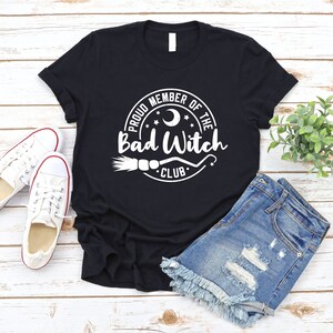 May include: Black t-shirt with a white circle graphic that says "Proud Member of the Bad Witch Club" with a crescent moon, stars, and a broom.