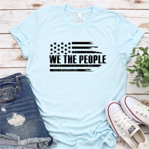 Op de afbeelding: Lichtblauw T-shirt met een zwart, verweerd ontwerp van de Amerikaanse vlag. De tekst "WE THE PEOPLE" staat eronder. De afbeelding toont een casual, patriottisch thema, gecombineerd met een spijkerbroek en witte sneakers.