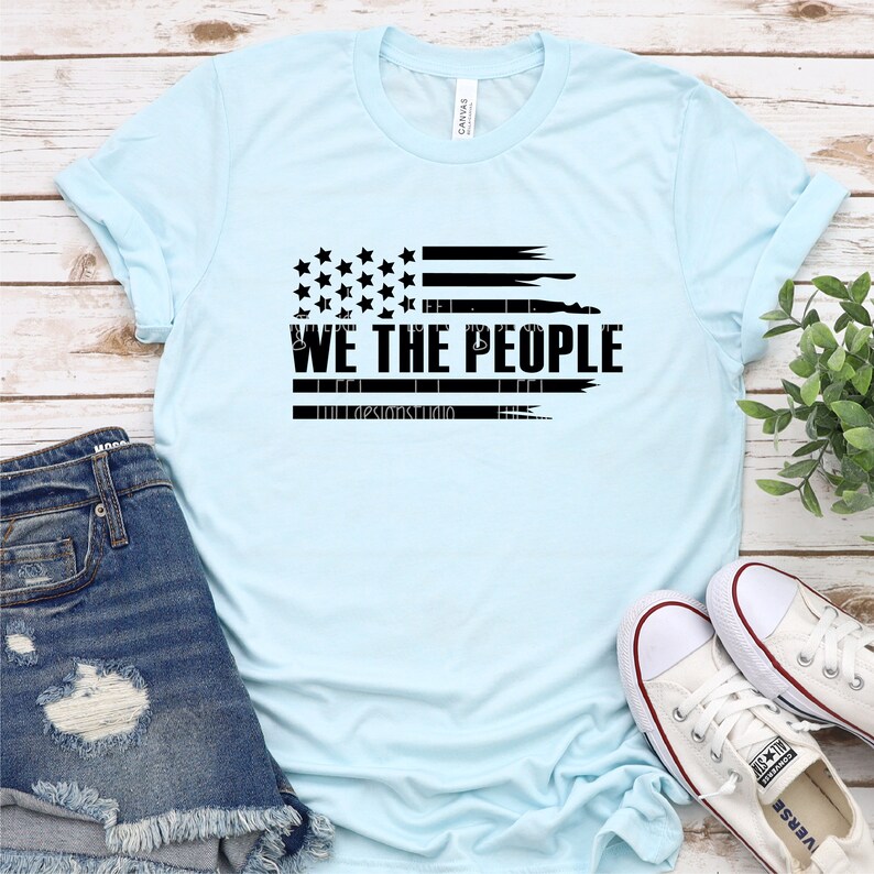 Op de afbeelding: Lichtblauw T-shirt met een zwart, verweerd ontwerp van de Amerikaanse vlag. De tekst "WE THE PEOPLE" staat eronder. De afbeelding toont een casual, patriottisch thema, gecombineerd met een spijkerbroek en witte sneakers.