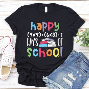 May include: Black t-shirt with the text "happy (9x9)+(6x3)+1 days of school!" in colorful letters. A stack of colorful books is in the middle of the text.