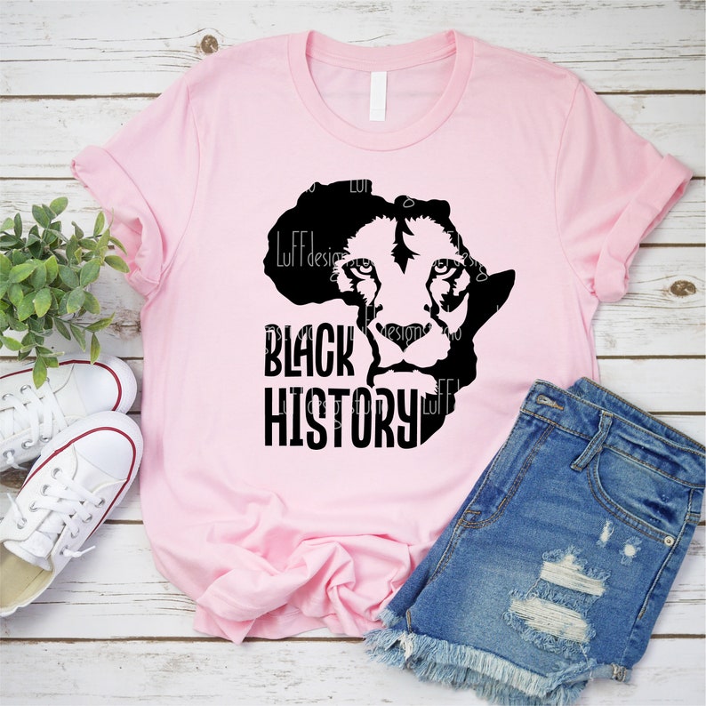 May include: A light pink t-shirt featuring a black graphic of a lion's face within the outline of the African continent. The words "BLACK HISTORY" are printed in bold, black letters below the graphic. The shirt is paired with denim shorts and white sneakers.