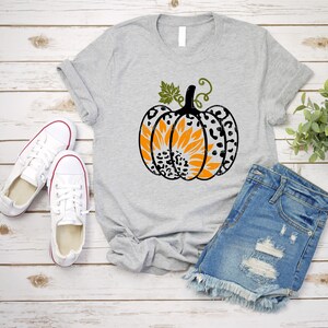 K&ouml;nnte beinhalten: Ein graues T-Shirt mit einem schwarzen und wei&szlig;en Leopardenmuster-K&uuml;rbis mit einer orangefarbenen Sonnenblume im Inneren. Der K&uuml;rbis hat einen gr&uuml;nen Stiel und Bl&auml;tter. Das T-Shirt liegt auf einer wei&szlig;en Holzoberfl&auml;che mit einem Paar wei&szlig;en Turnschuhen und einer blauen Jeans-Shorts.