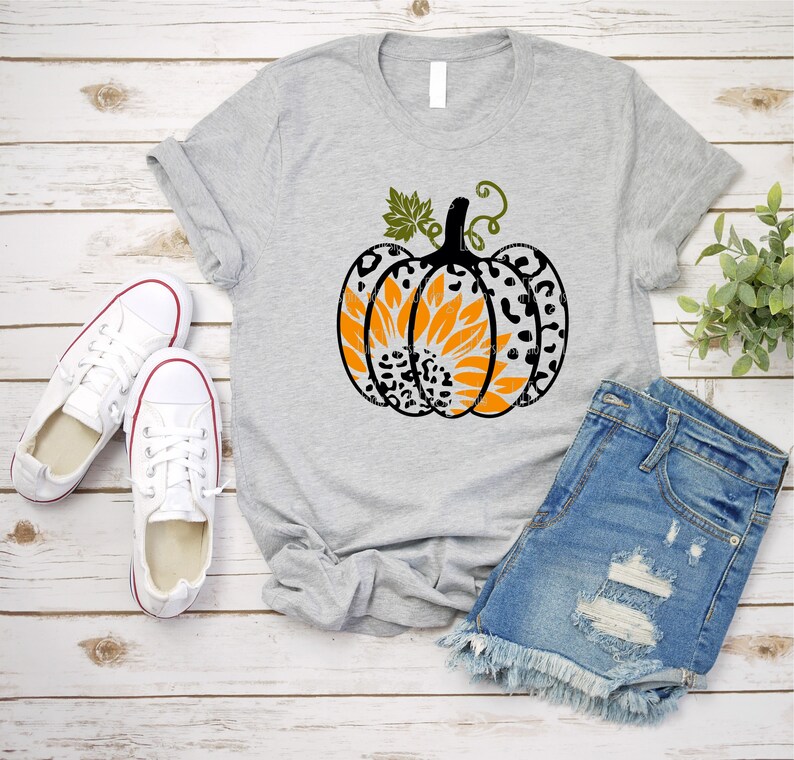 K&ouml;nnte beinhalten: Ein graues T-Shirt mit einem schwarzen und wei&szlig;en Leopardenmuster-K&uuml;rbis mit einer orangefarbenen Sonnenblume im Inneren. Der K&uuml;rbis hat einen gr&uuml;nen Stiel und Bl&auml;tter. Das T-Shirt liegt auf einer wei&szlig;en Holzoberfl&auml;che mit einem Paar wei&szlig;en Turnschuhen und einer blauen Jeans-Shorts.