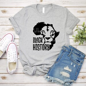 May include: A light gray t-shirt featuring a black graphic of a lion's face within the outline of the African continent. The words "BLACK HISTORY" are printed in bold, black letters below the lion. White sneakers and denim shorts are also visible.