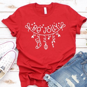 Op de afbeelding: Rood T-shirt met een witte afbeelding van een kerstkrans met de tekst "Be Jolly" die eraan hangt. De krans heeft een sok, een ster, een snoepstok en een kerstbal.