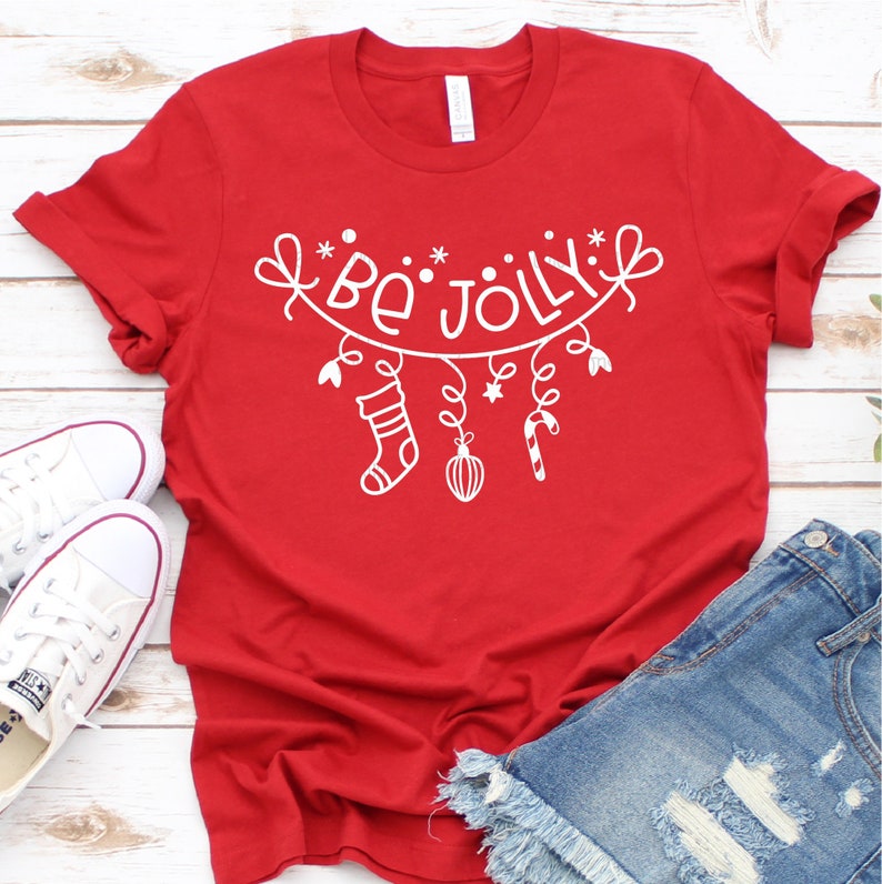 Op de afbeelding: Rood T-shirt met een witte afbeelding van een kerstkrans met de tekst "Be Jolly" die eraan hangt. De krans heeft een sok, een ster, een snoepstok en een kerstbal.