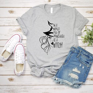 Peut inclure: Un t-shirt gris avec un graphique noir d'une sorci&egrave;re avec le texte "Dans un monde plein de princesses, sois une sorci&egrave;re".